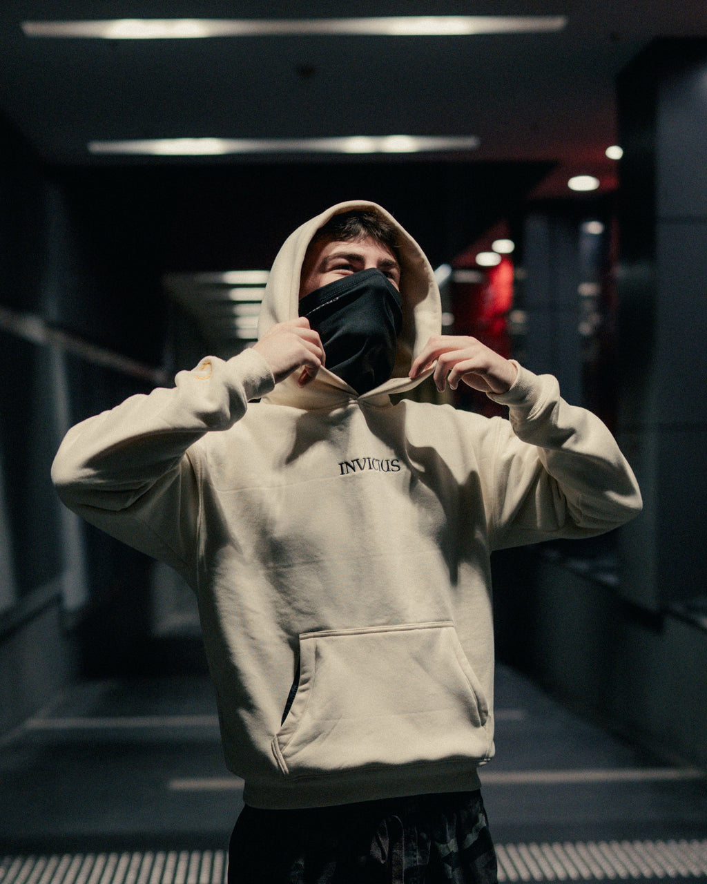 Person wearing an off-white hoodie with 'INVICTUS' branding in a dimly lit indoor setting.