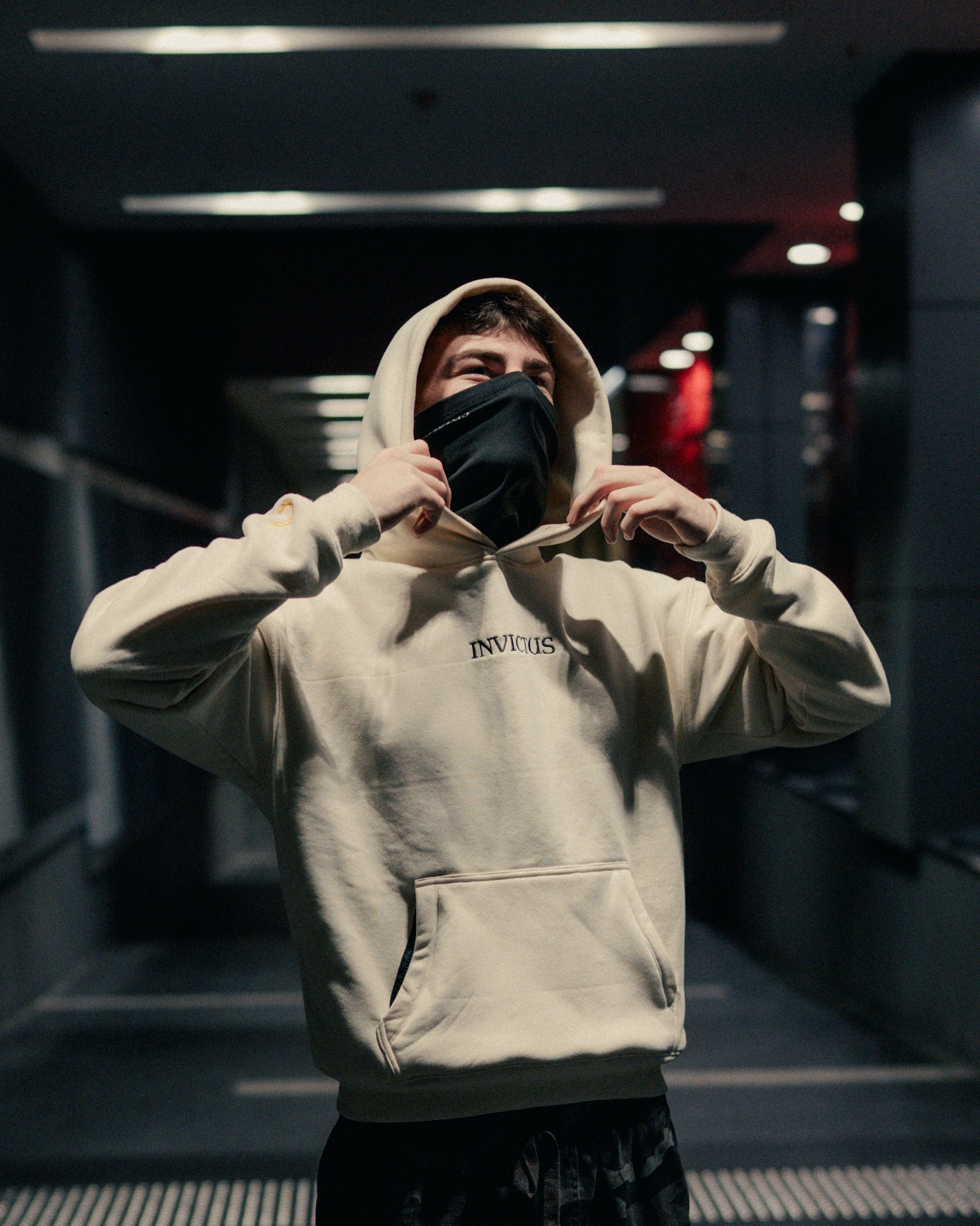 Person wearing an off-white hoodie with 'INVICTUS' branding in a dimly lit indoor setting.